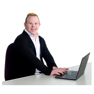 A person sitting at a desk, smiling, and working on a laptop.